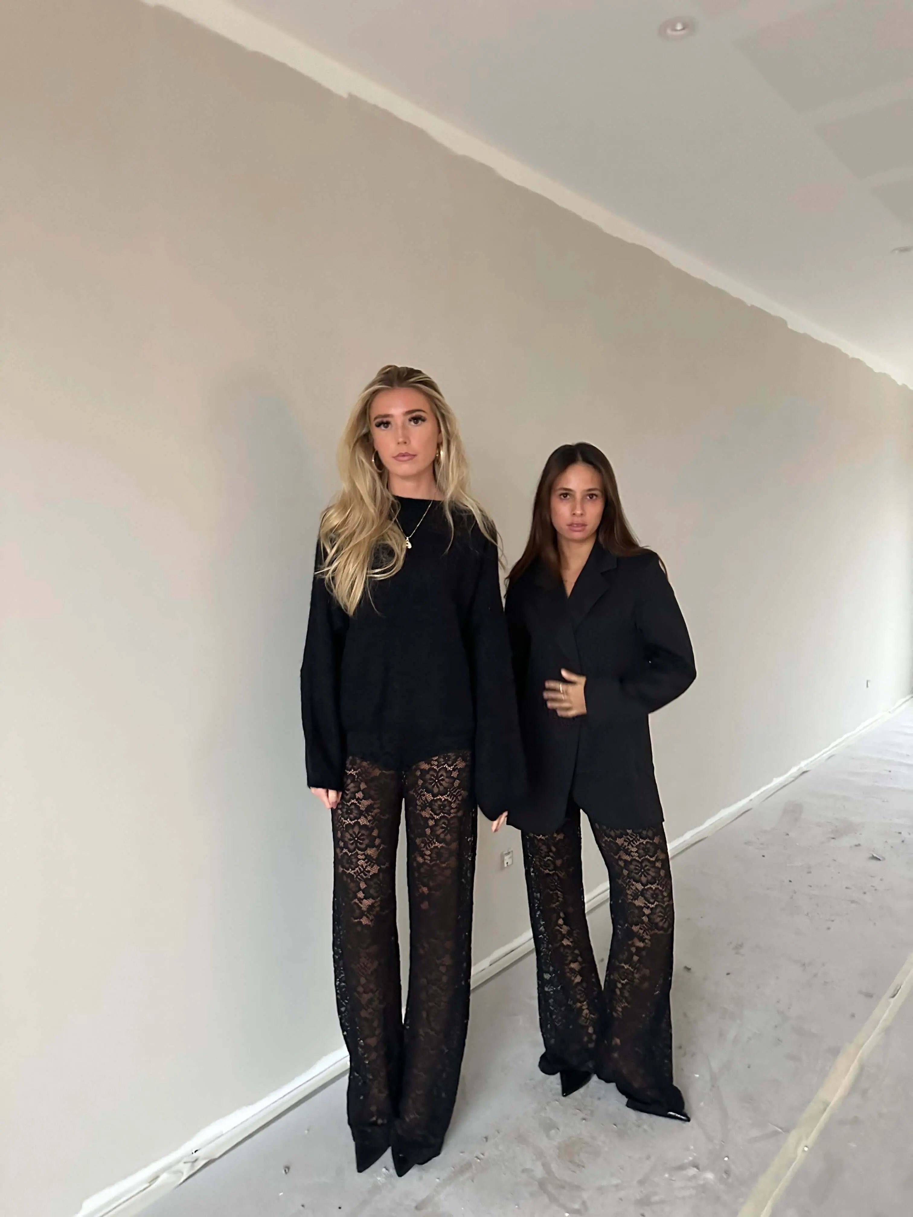 Two women wearing black tops and Valentina™ Luxe Kanten Broek with lace details in a neutral-toned room.